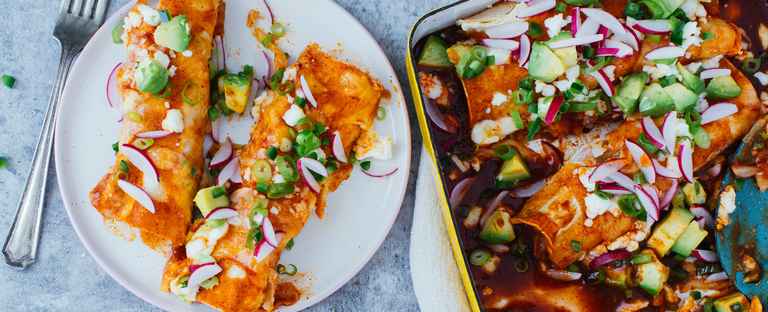 Two enchiladas on a plate next to a baking dish with enchiladas